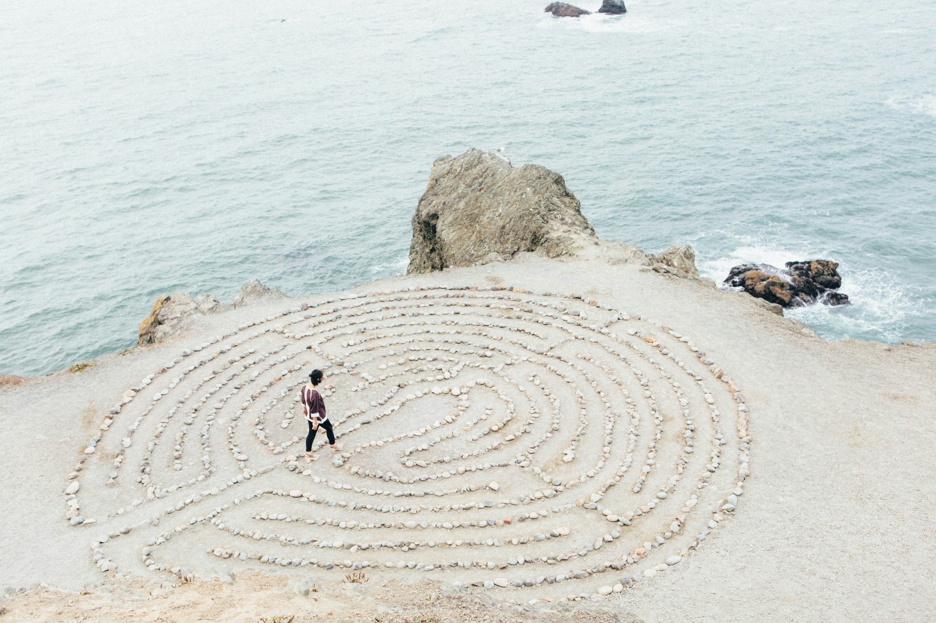 A solitary figure walking a stone labyrinth above the sea — a contemplative practice
