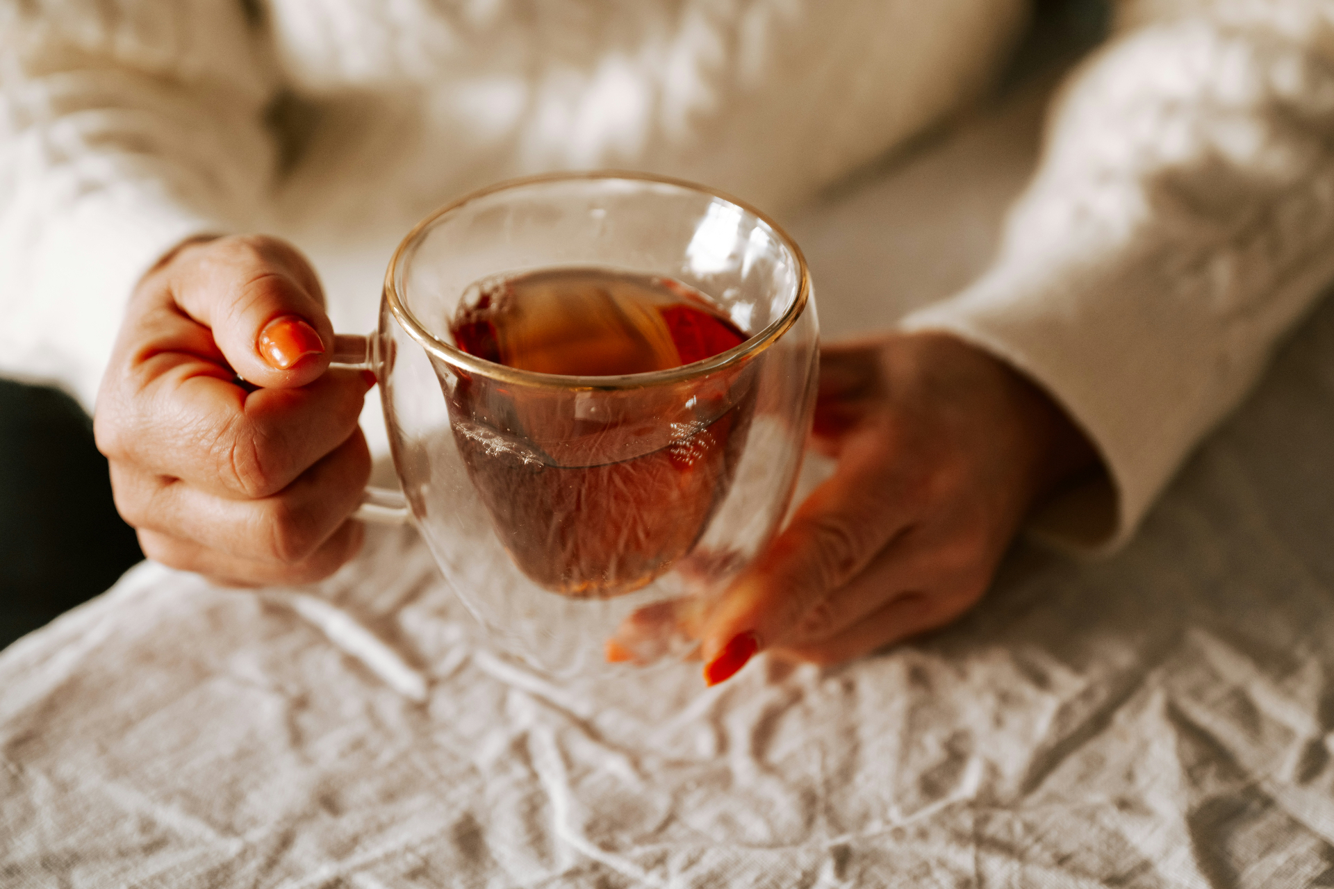 Hands cradling a clear glass mug of warm tea on soft linen